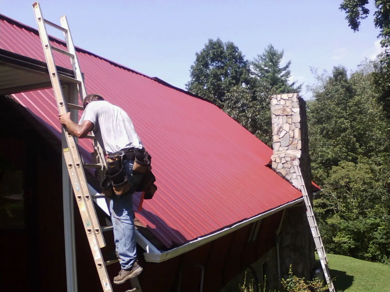 Roofer on a ladder beside a red metal roof and stone chimney — Soffit & Fascia Repair by Roofing Brothers in Ahsahka